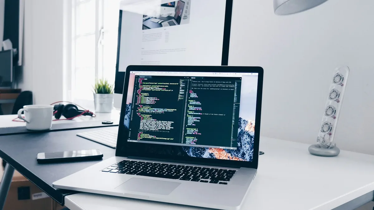 A laptop showing a code editor with CSS on a clean desk beside an external monitor — a frontend developer's workspace